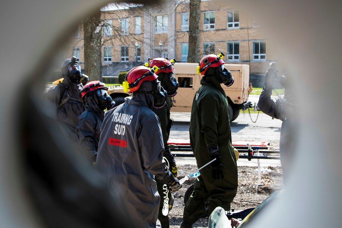 Soldiers wearing gas masks, hard hats and other protective gear walk near a building.