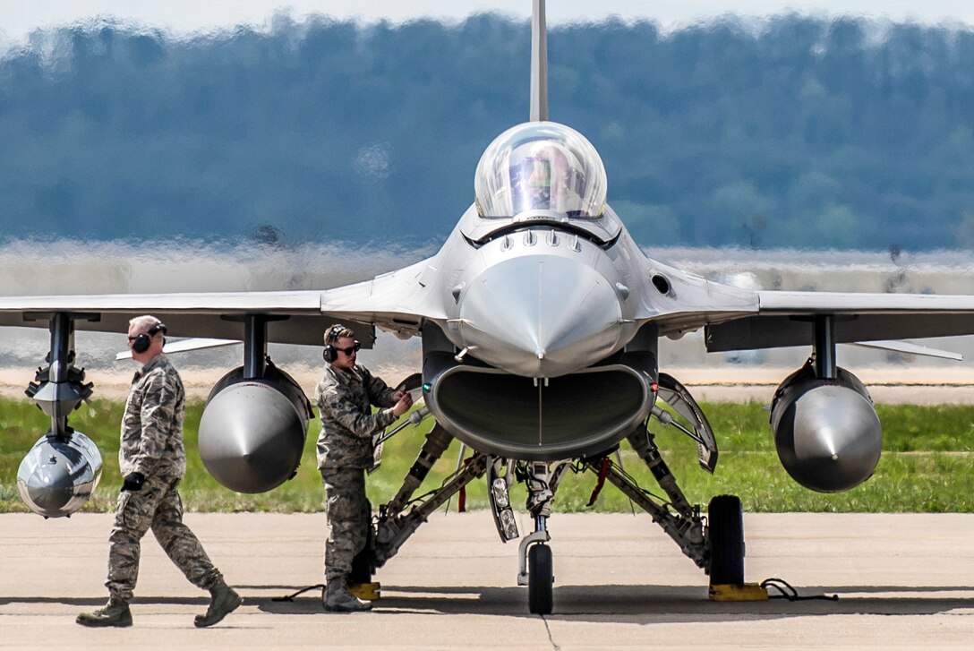 An airman works on a jet on a flightline as another airman walks nearby.