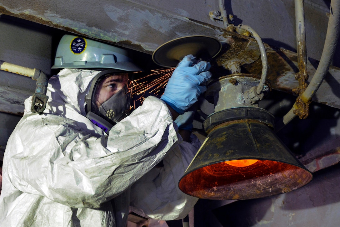 A sailor in white coveralls and a hardhat uses a sander on an overhead structure.