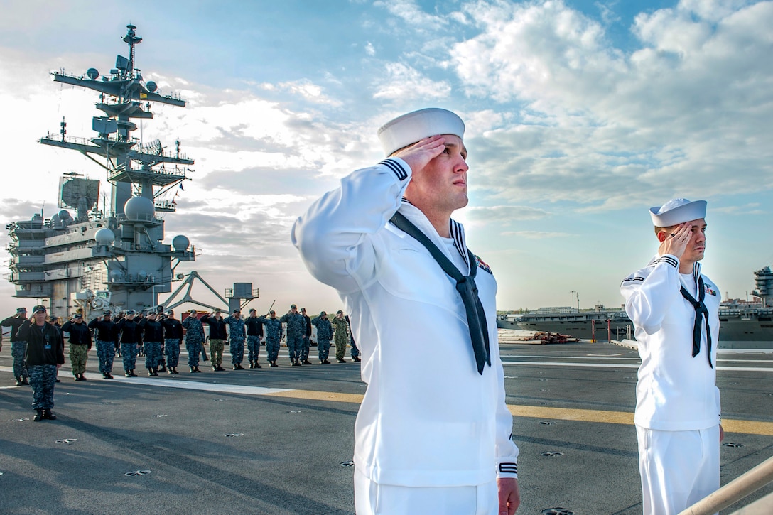 Two sailors salute on a ship's flight deck, as others gather behind them in the distance.