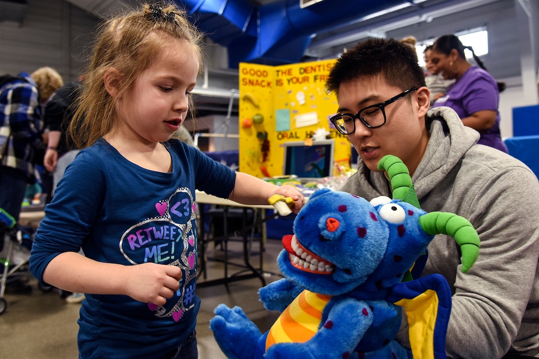 A girl brushes a blue dragon stuffed animal's teeth as an airman observes.