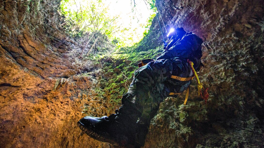 A Marine in protective gear hangs from a wire inside a cave as light shines in from the opening.