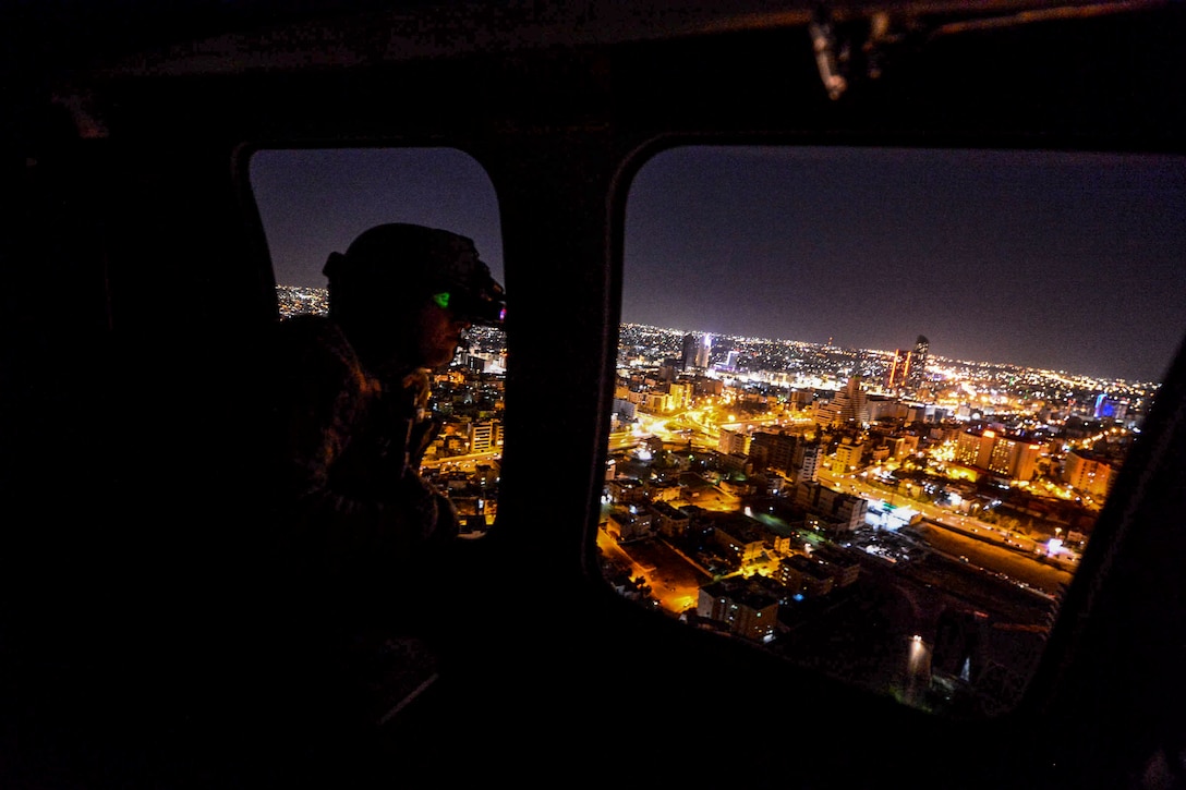 A soldier looks out from a window of an in-flight helicopter at city lights.