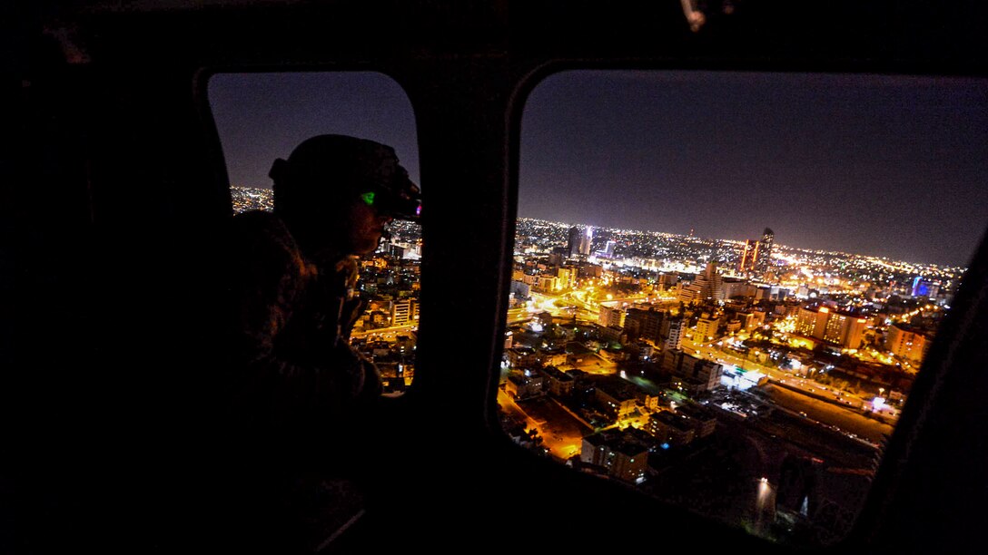 A soldier looks out from a window of an in-flight helicopter at city lights.