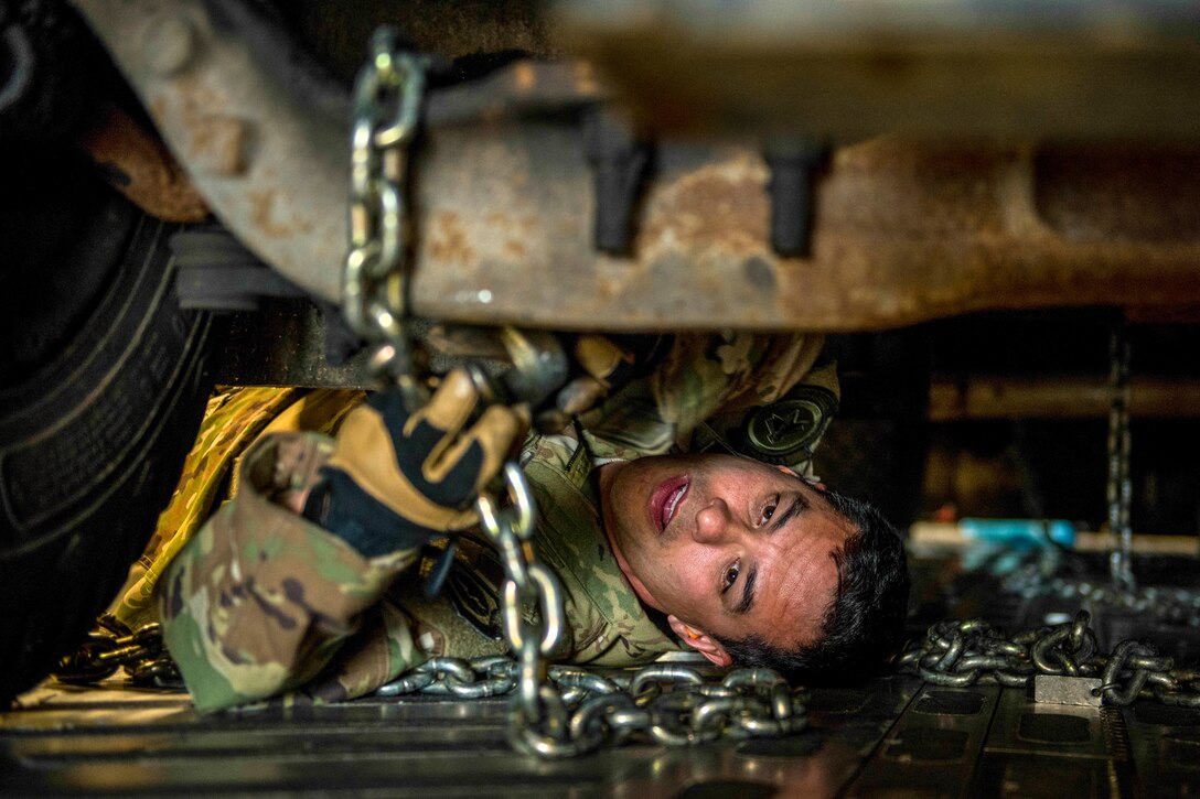 An airman lies on an aircraft floor and manipulates a chain attached to a vehicle