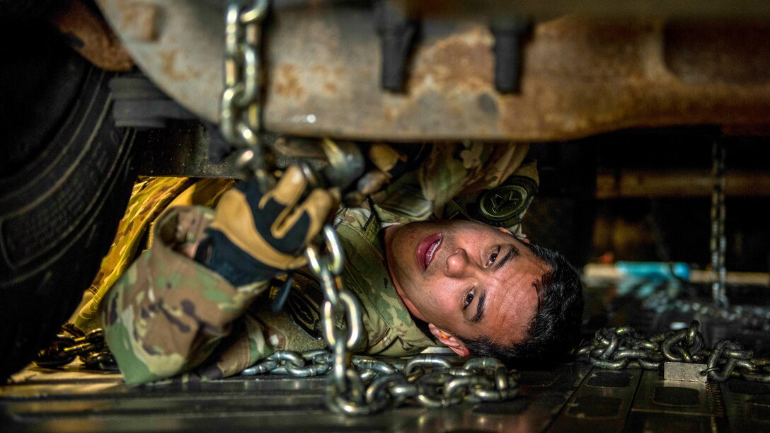 An airman lies on an aircraft floor and manipulates a chain attached to a vehicle