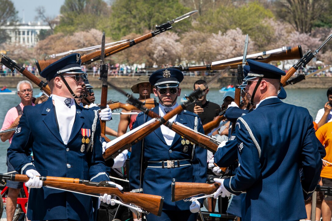 Air Force honor guard members toss rifles in formation as a crowd watches against a background of blossoming cherry trees.
