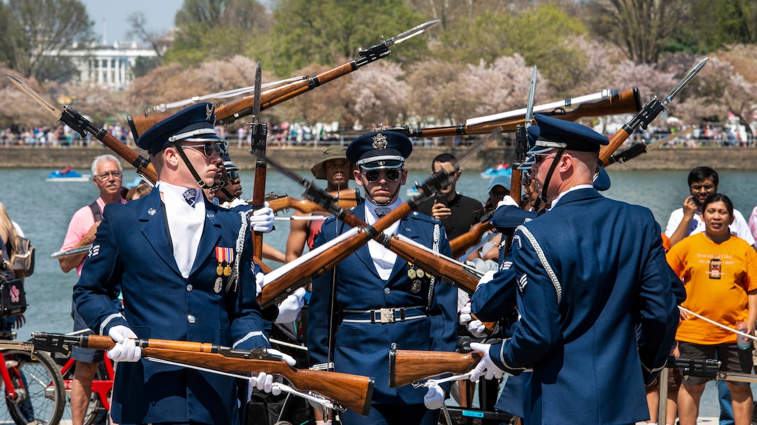 Air Force honor guard members toss rifles in formation as a crowd watches against a background of blossoming cherry trees.