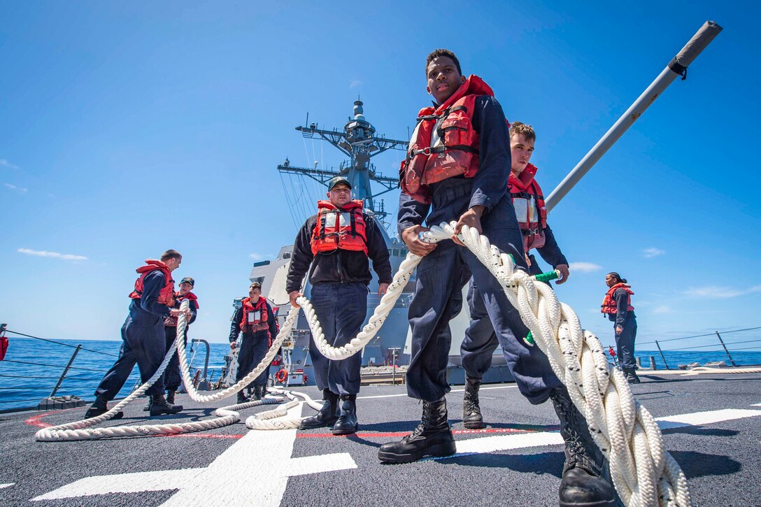 Sailors in red life vests handle a rope aboard a ship.