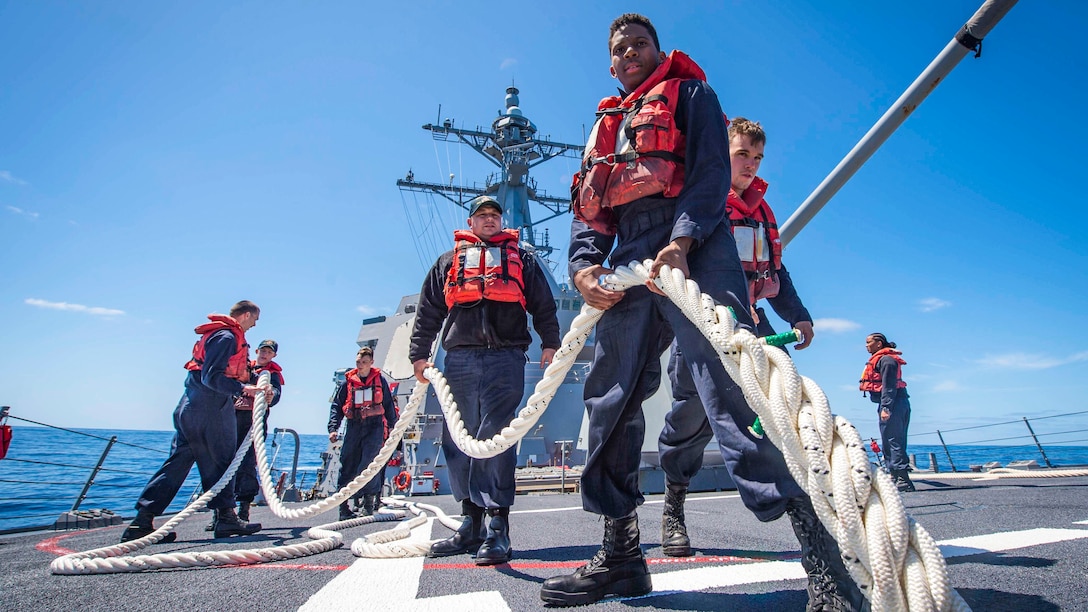 Sailors in red life vests handle a rope aboard a ship.