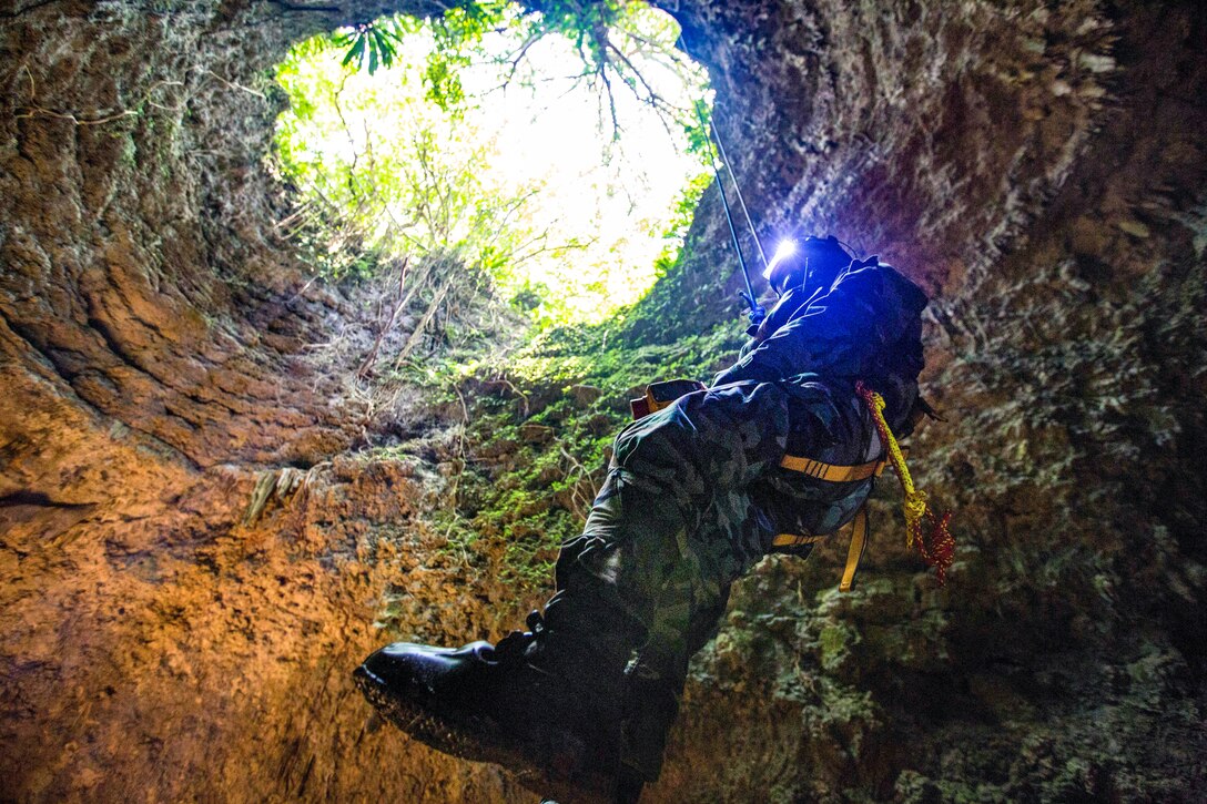 A Marine in protective gear hangs from a wire inside a cave as light shines in from the opening.