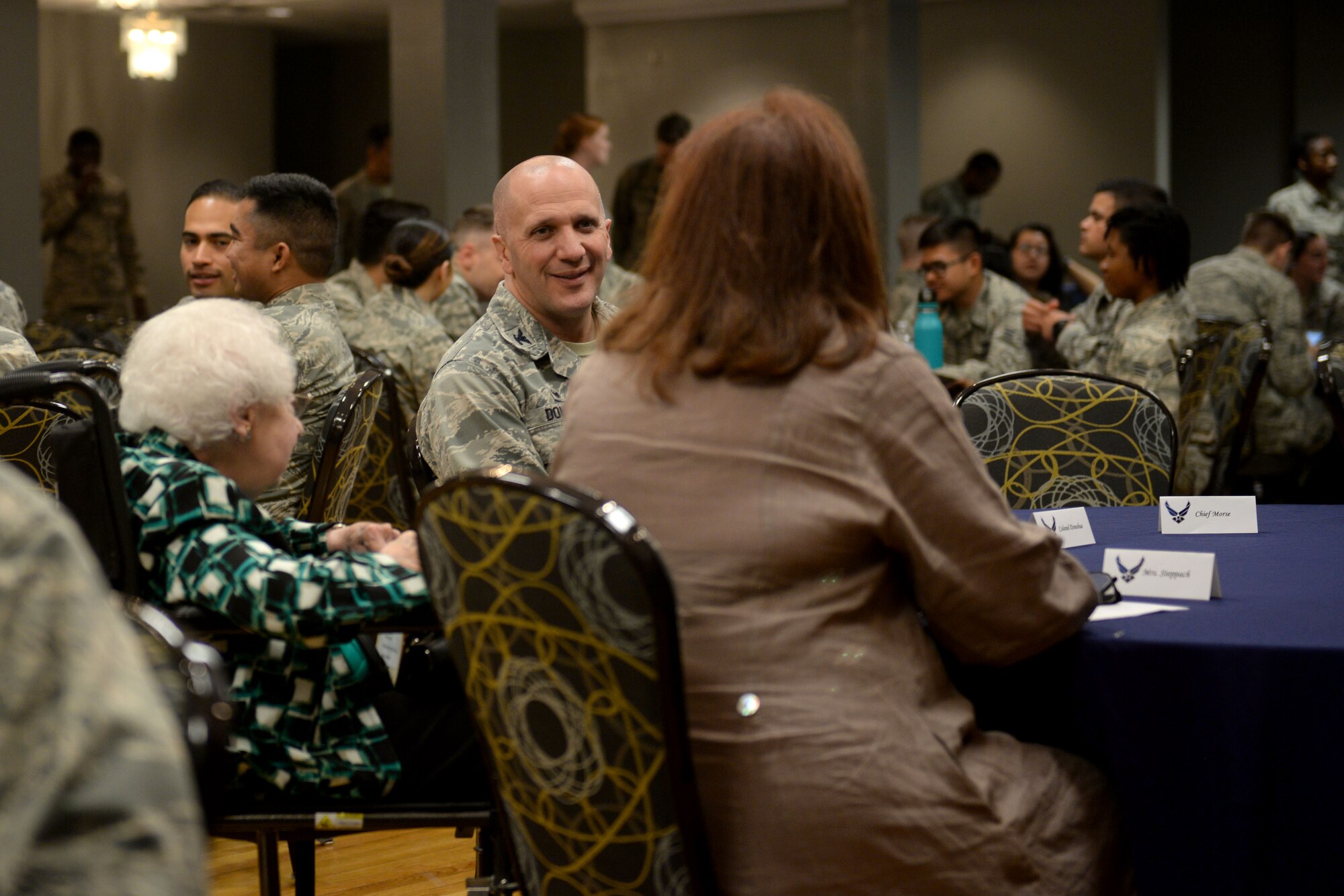 A man in the Airman Battle Uniform smiles at a woman with red hair wearing brown shawl and an elderly woman in a blue, white and black shirt.