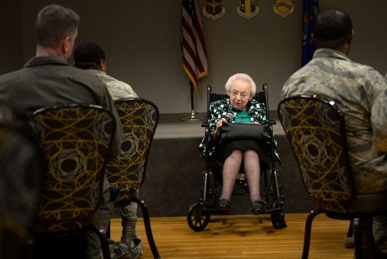 An elderly woman in a wheel chair wearing a blue, white and black shirt holds a microphone up to her mouth in front of numerous people in the Airman Battle Uniform.