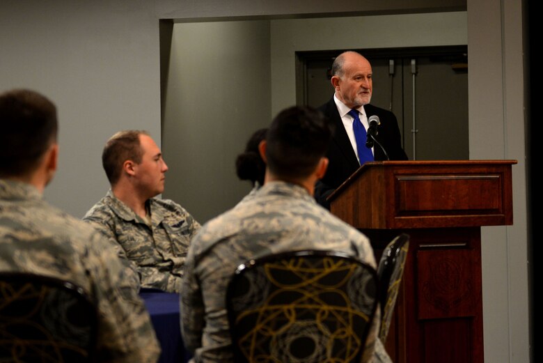 A man wearing a black suit with a white undershirt and a blue tie stands at a podium speaking into a microphone while people in the Airman Battle Uniform look at him while sitting at tables.