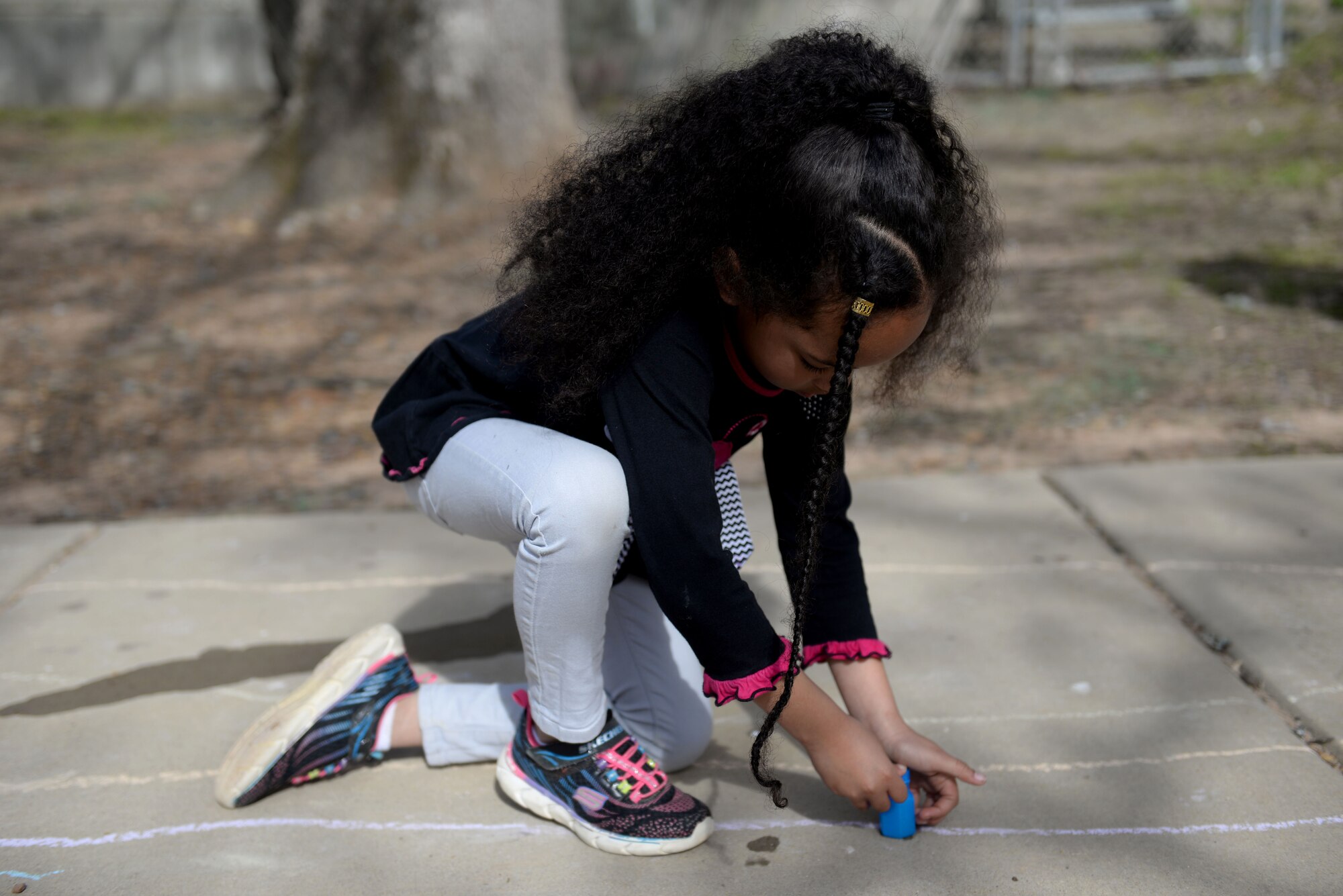 A  young child with a long black braid in the front of her head and long black hair pulled into a ponytail puts together a blue bottle on the ground.