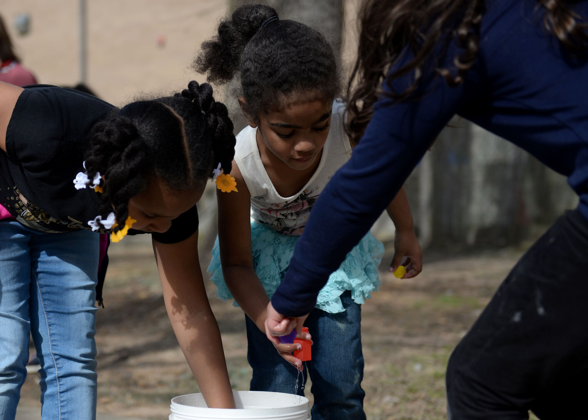 Three young girls with black hair reach into a white bucket to fill small colored bottles with water.