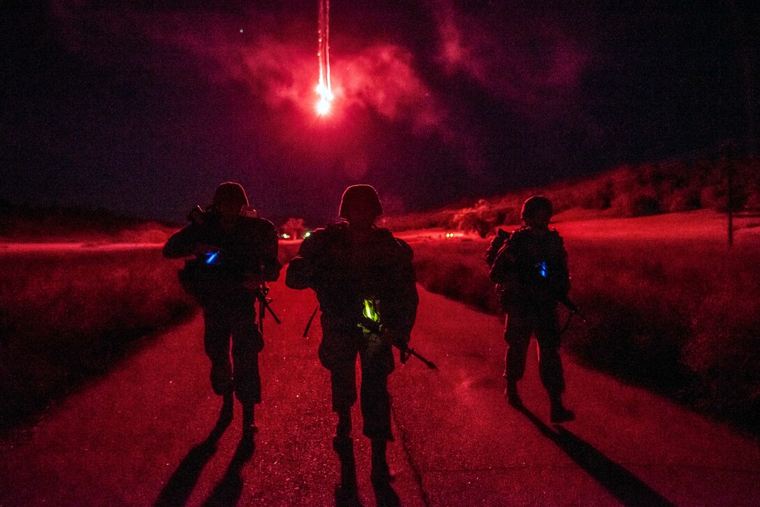 Red fireworks illuminate a night sky as three soldiers, shown in silhouette, walk with weapons on a roadway.