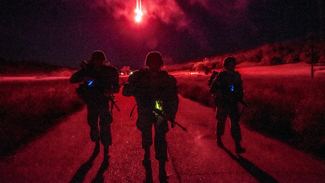 Red fireworks illuminate a night sky as three soldiers, shown in silhouette, walk with weapons on a roadway.