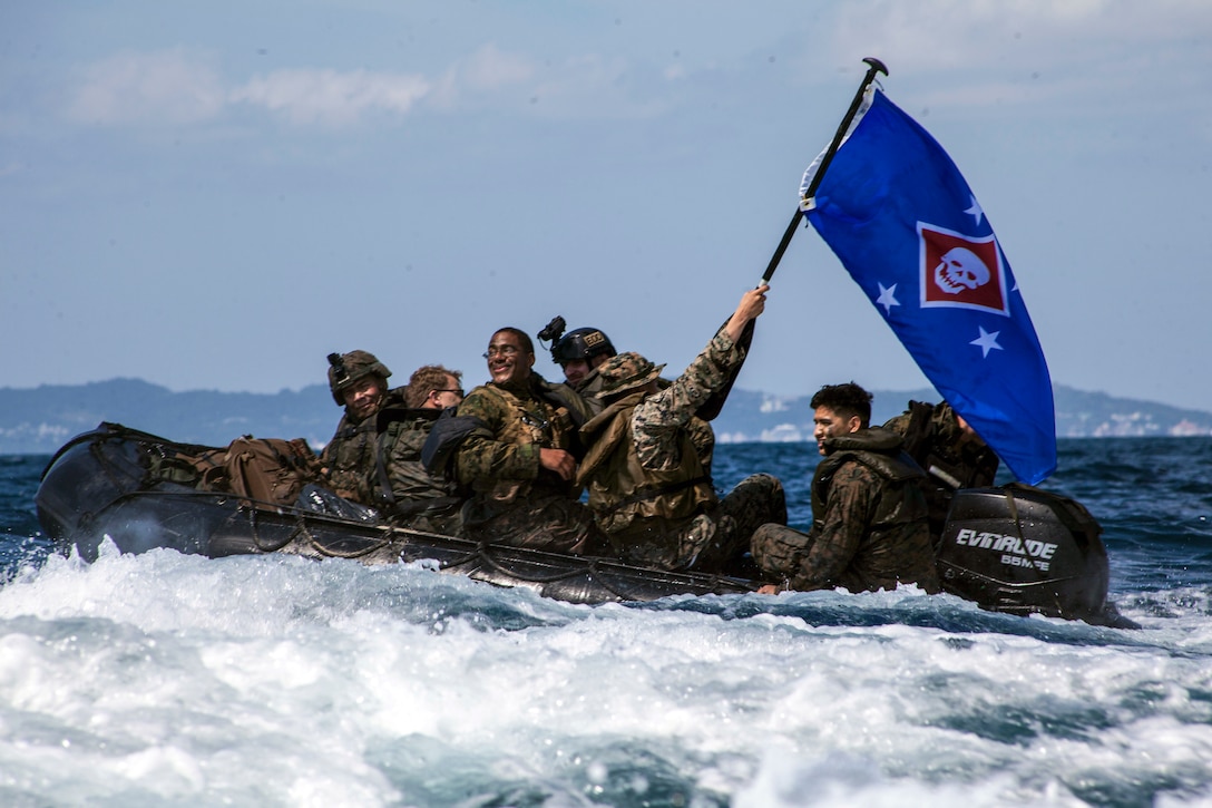 Marines ride aboard a rubber boat in white waters, with one hoisting up a blue flag with a skull emblem.