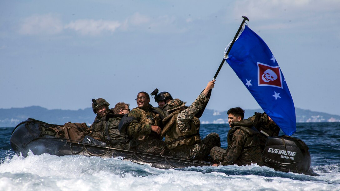 Marines ride aboard a rubber boat in white waters, with one hoisting up a blue flag with a skull emblem.