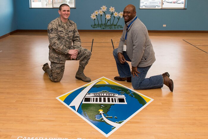 Two men kneel on a floor next to a painted logo.