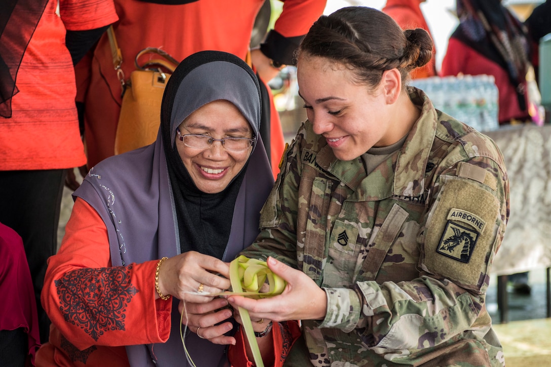 A smiling soldier holds strips of leaves as a smiling Malaysian woman helps her weave them.