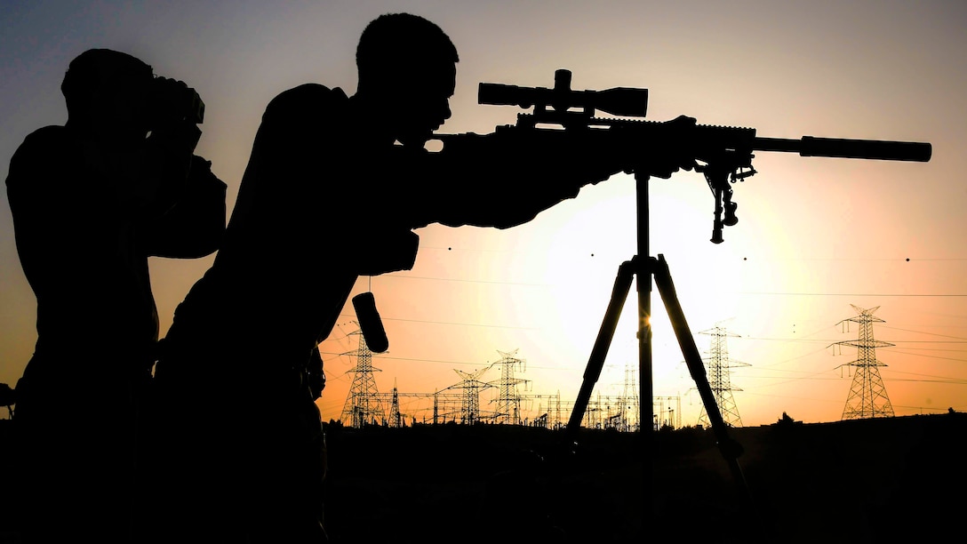 A Marine, shown in silhouette, looks through a sniper rifle scope, as someone looks through binoculars beside him.