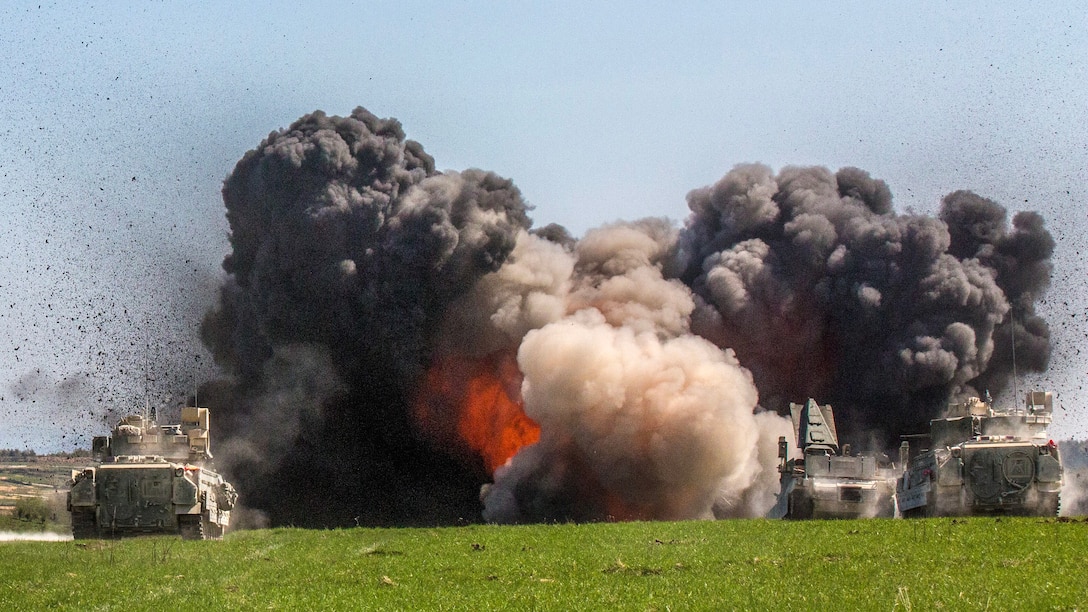 Black clouds of smoke and flames fill the air in a field, as tanks conduct training.
