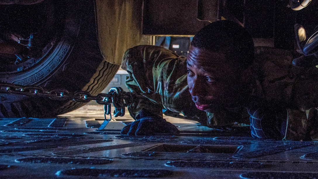 An airman lies on his stomach and looks at the undercarriage of a vehicle.