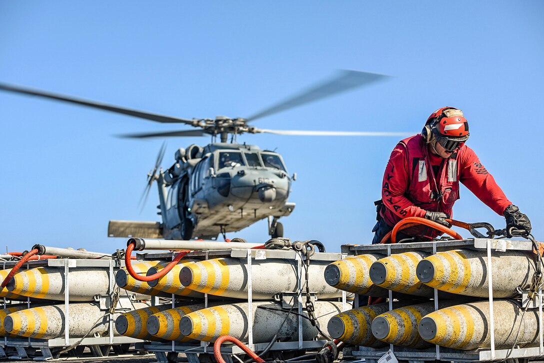 A sailor in red hooks a cable to a pallet of ordnance as a helicopter flies behind him.