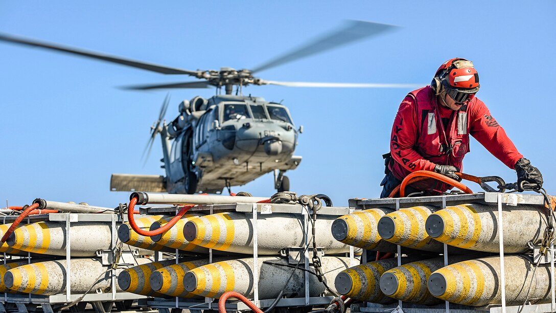 A sailor in red hooks a cable to a pallet of ordnance as a helicopter flies behind him.