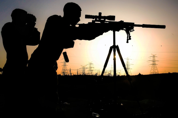 A Marine, shown in silhouette, looks through a sniper rifle scope, as someone looks through binoculars beside him.
