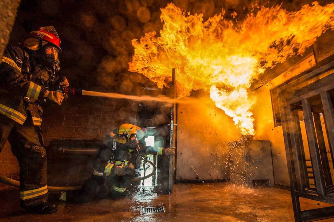 An airman sprays flames with a hose inside a room, as another firefighter observes.