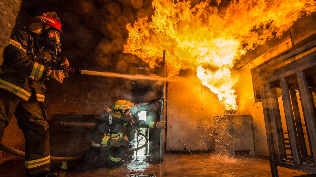 An airman sprays flames with a hose inside a room, as another firefighter observes.