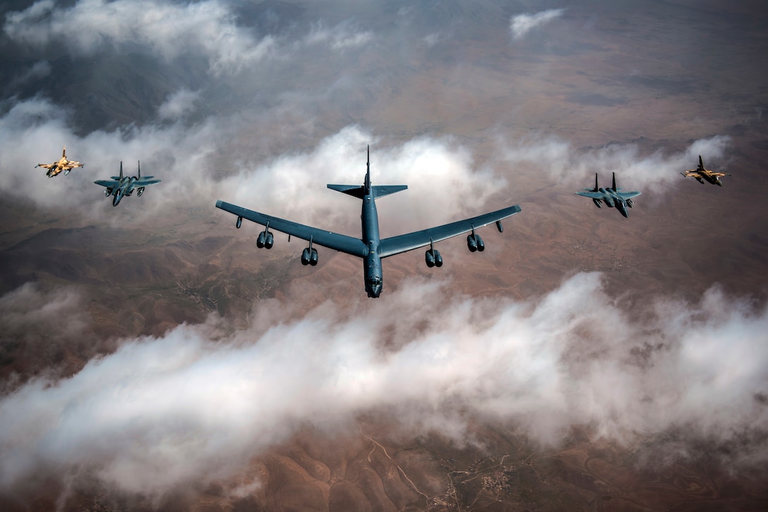 Five aircraft fly in formation over clouds and brown terrain.