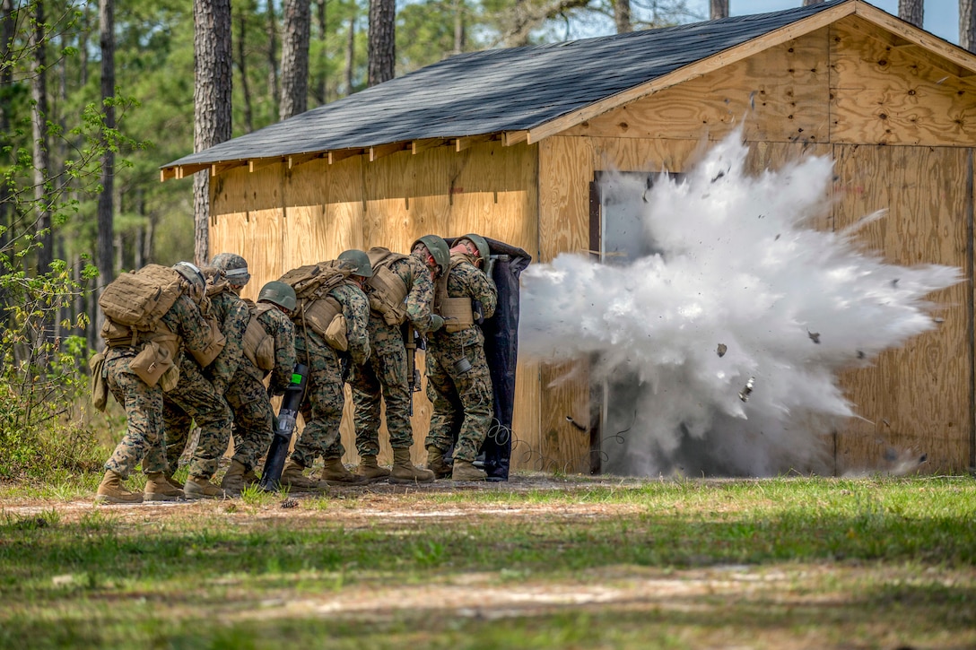 Marines crouch in a line by a makeshift building as a charge explodes, creating plumes of white smoke.