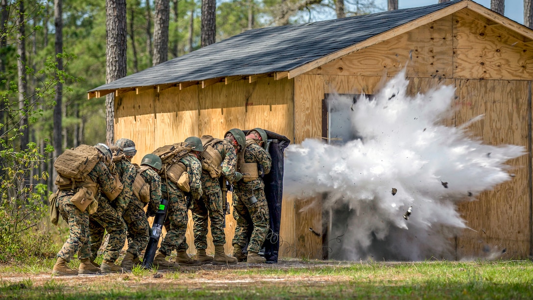 Marines crouch in a line by a makeshift building as a charge explodes, creating plumes of white smoke.