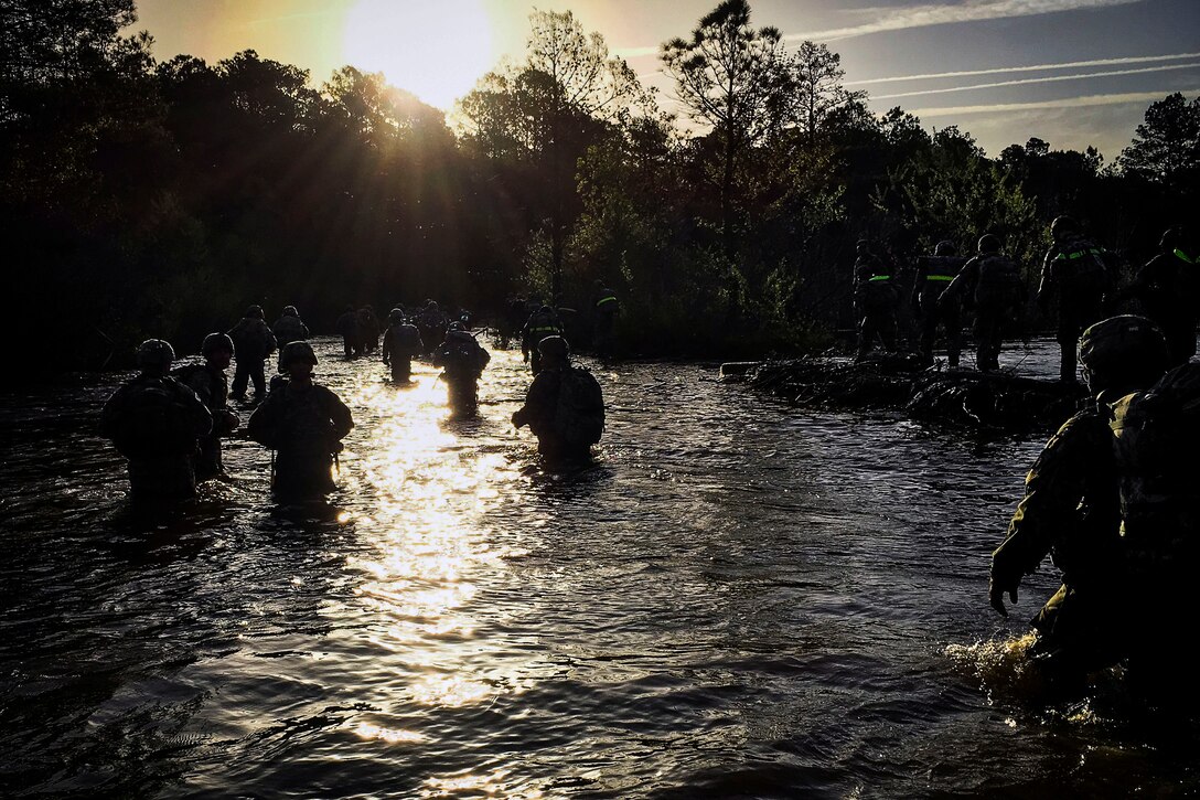 Soldiers, shown in silhouette, wade through thigh-high waters at either sunrise or sunset.