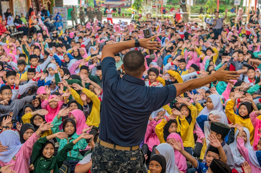 A sailor, shown from behind, holds his hands above his head and waves as an audience of children does the same.