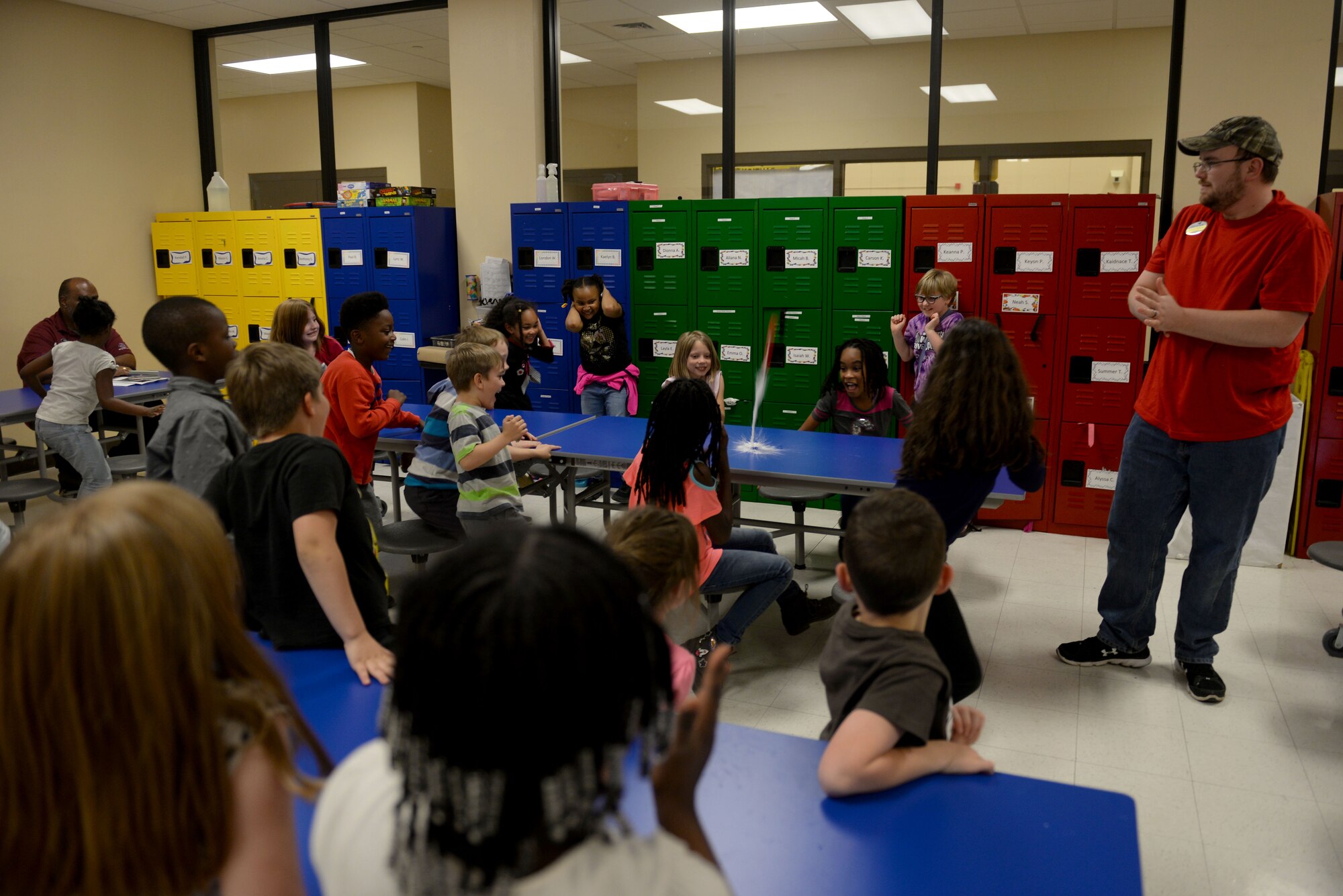 Numerous children sit around blue tables with a man in a red shirt demonstrates an educational bottle rocket fueled with water and baking soda.