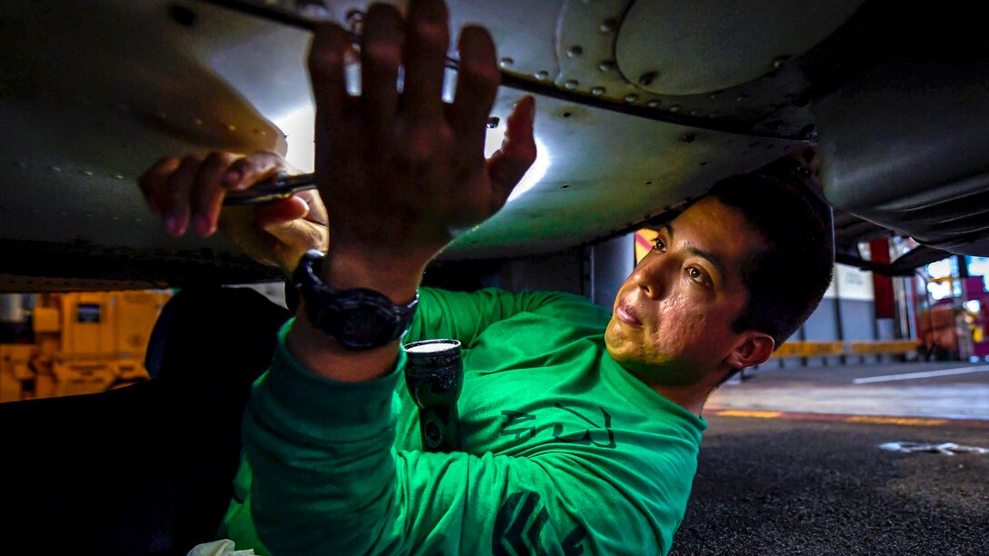 A sailor wearing a green shirt props a flashlight in his arm while lying on his back and working under an aircraft.