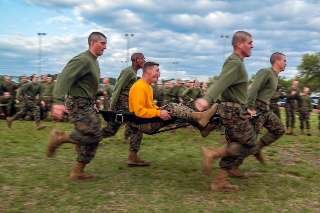 Marines carrying a fellow Marine on a stretcher run on a field as a crowd of Marines watch and cheer.