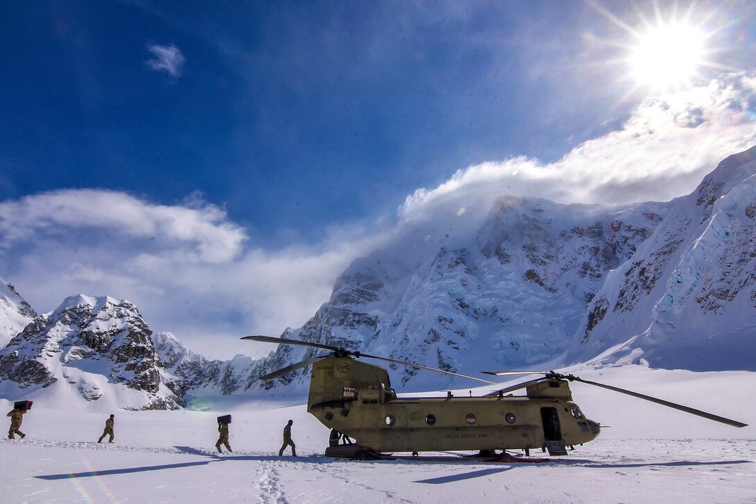 Soldiers walk across a field of snow toward a helicopter, with snowy mountain peaks in the background.