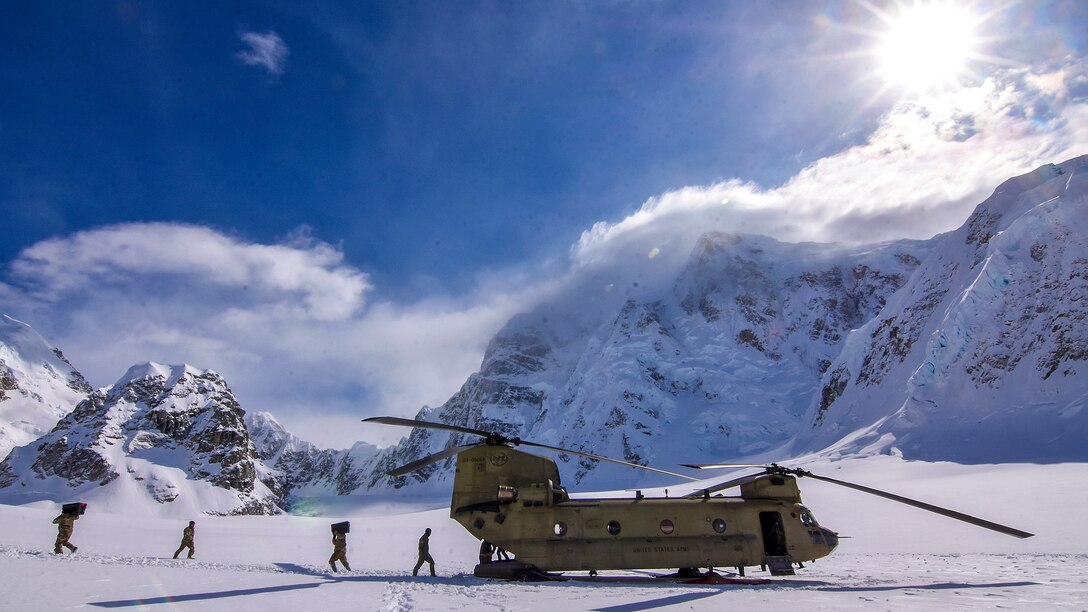Sailors walk across a field of snow toward a helicopter, with snowy mountain peaks in the background.