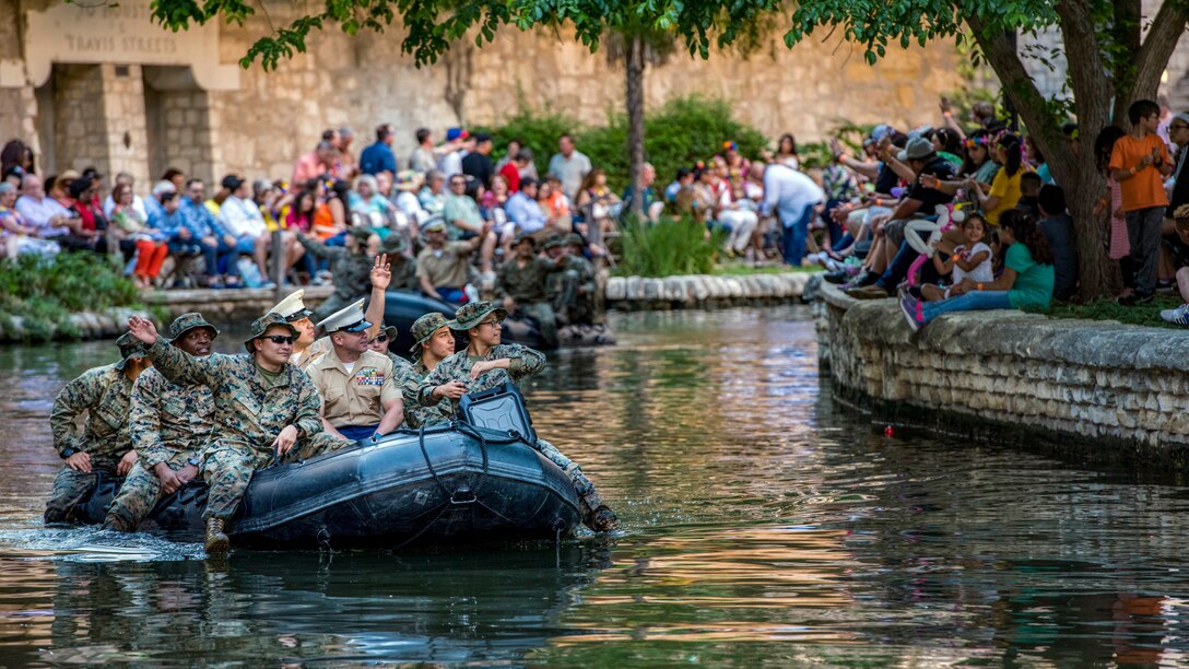 Marines float in a rubber boat down a river and wave at celebrants gathered on the banks.