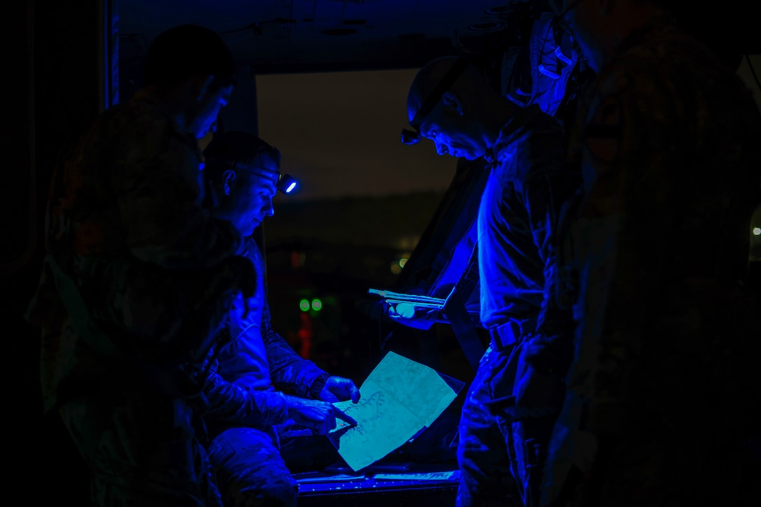 Three soldiers, illuminated by blue light, review a map and notes inside an aircraft or vehicle.