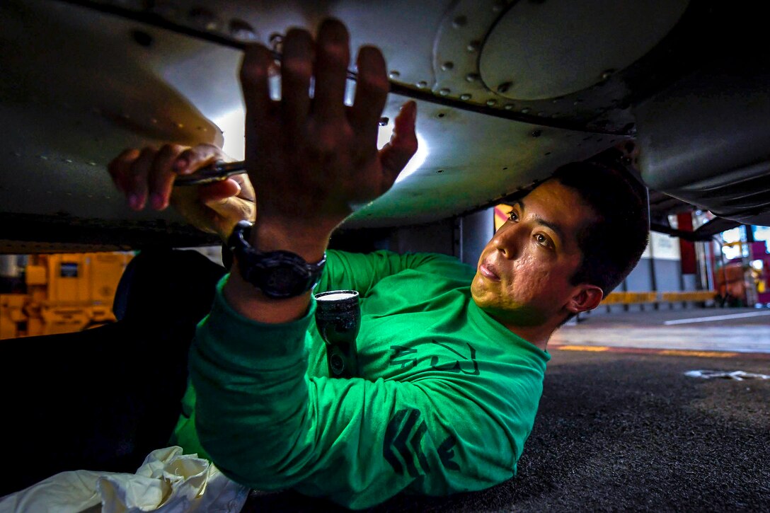 A sailor wearing a green shirt props a flashlight in his arm while lying on his back and working under an aircraft.