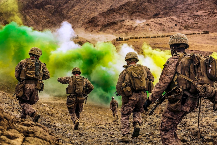 Marines run down a hill toward green smoke.