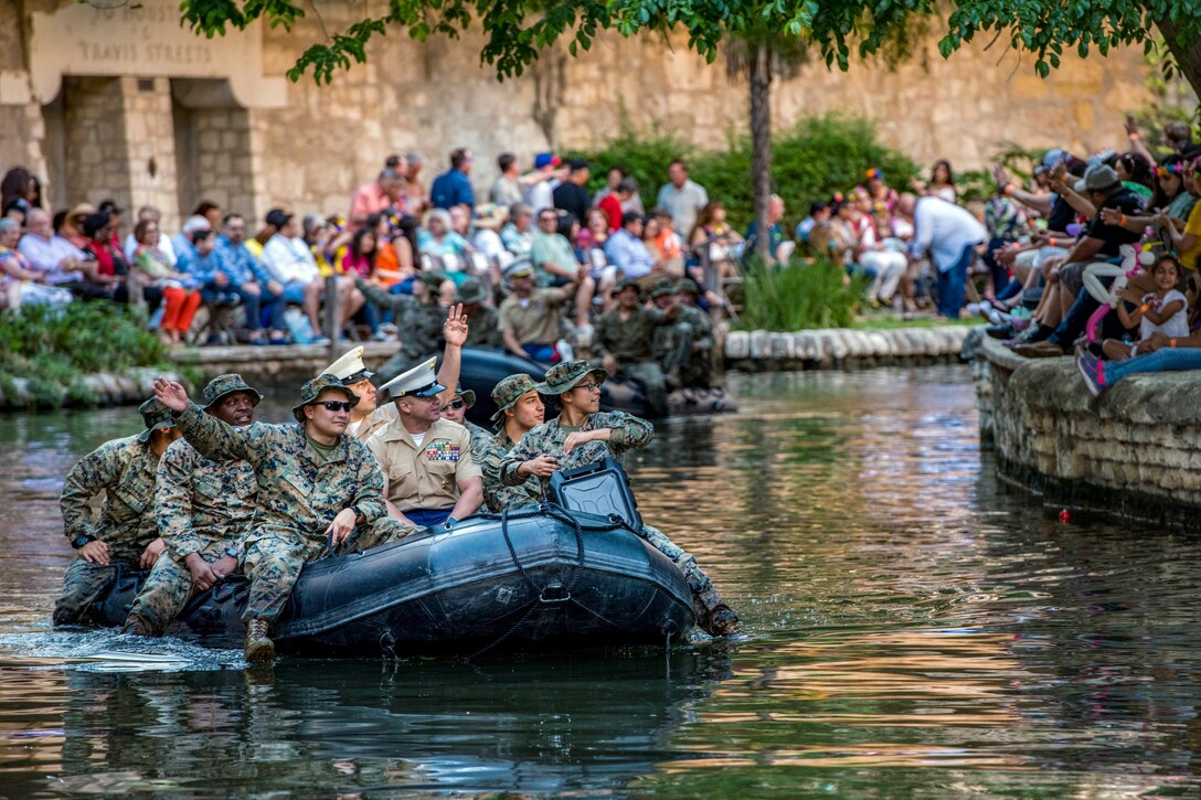 Marines float in a rubber boat down a river and wave at celebrants gathered on the banks.