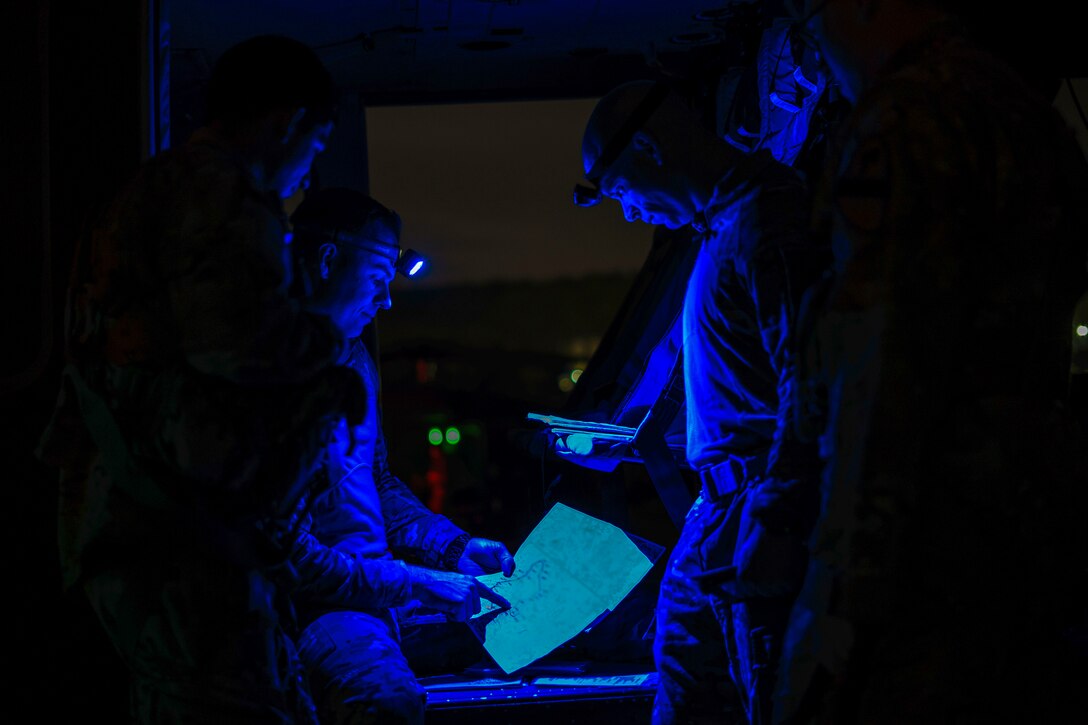 Three soldiers, illuminated by blue light, review a map and notes inside an aircraft or vehicle.